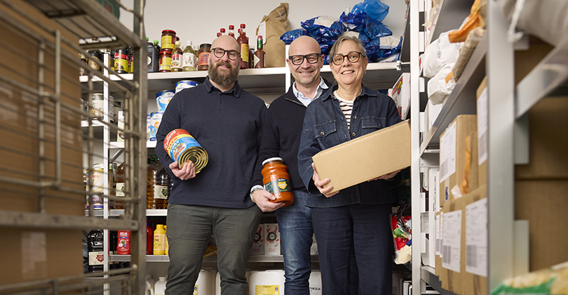 Tre personer står inne i ett lager med hyllor fyllda med kartonger och burkar.