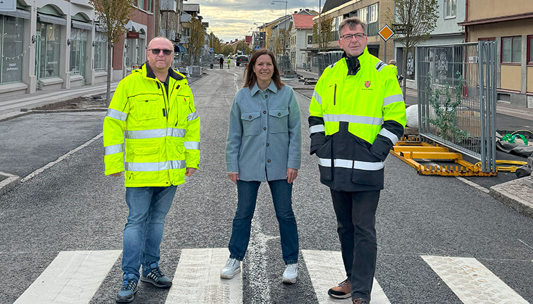 tre personer står på ett övergångsställe mitt i torggatan och blickar framåt. två personer är klädda i varselkläder och personen i mitten är vardagsklädd