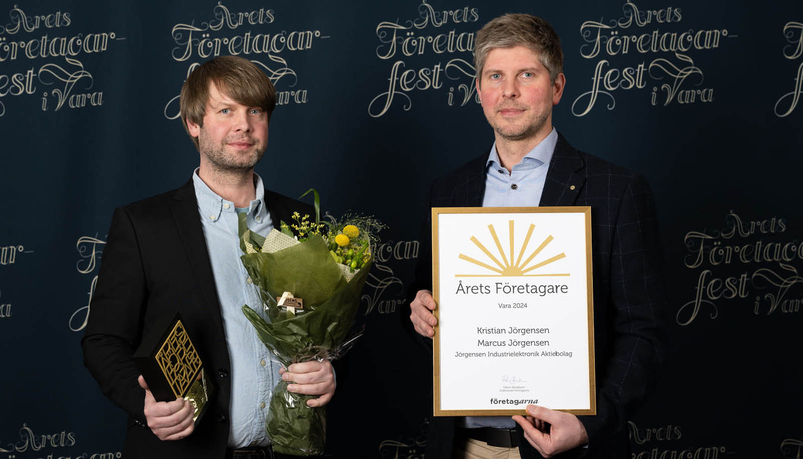 Två män i kostym, den ena håller i ett diplom och den andra i en blombukett.