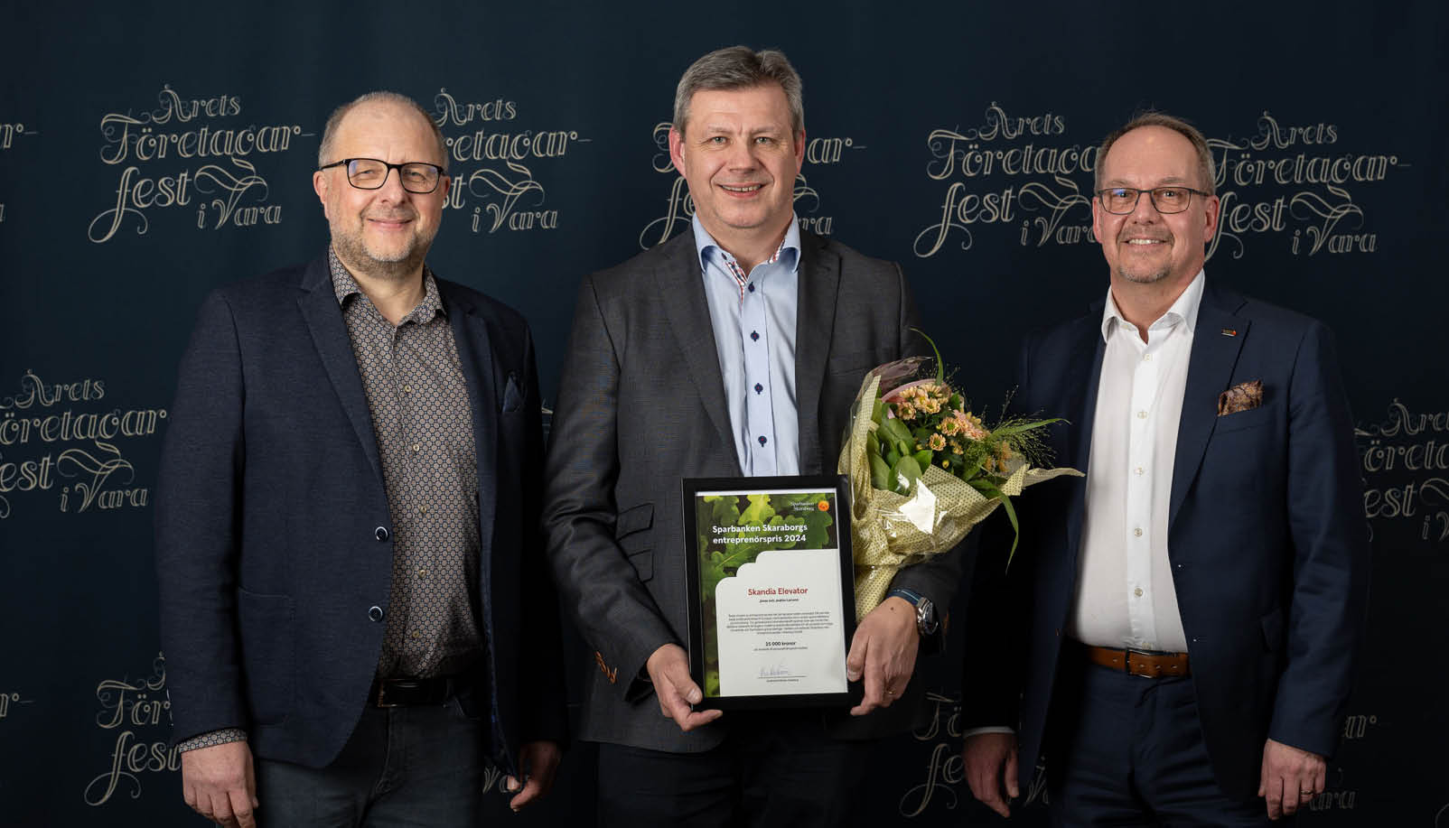 Tre män i kostym, den ena håller i ett diplom och en blombukett.