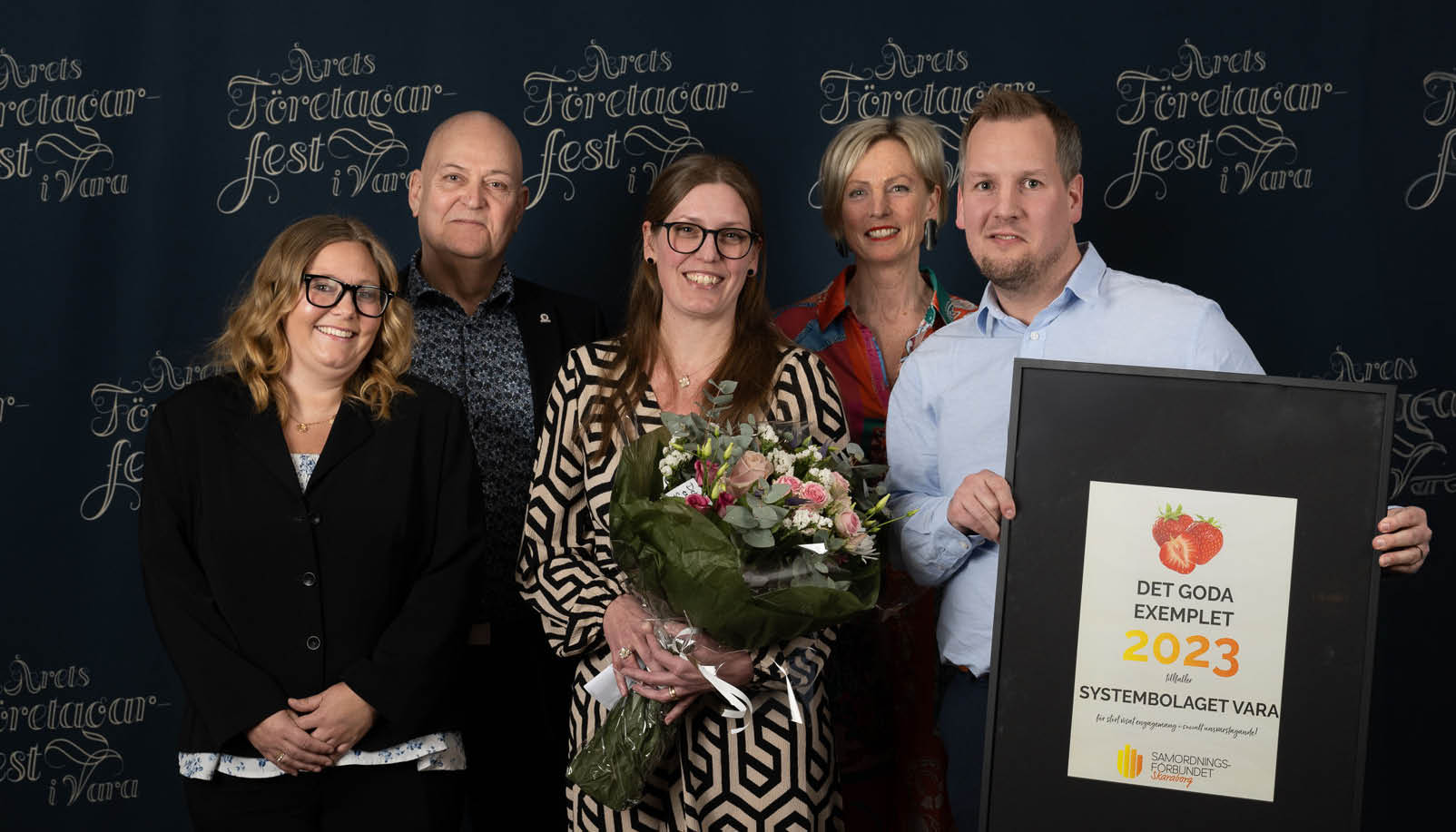 Tre kvinnor och två män i kostym, en man håller i ett diplom och en kvinna i en blombukett.