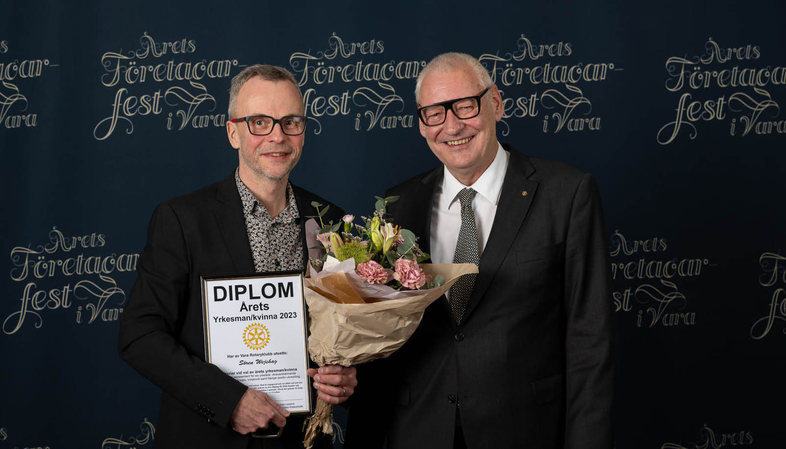 Två män i kostym, den ena håller i ett diplom och en blombukett.