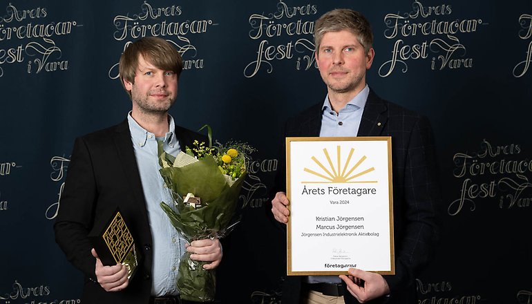 Två män i kostym, den ena håller i ett diplom och den andra i en blombukett.